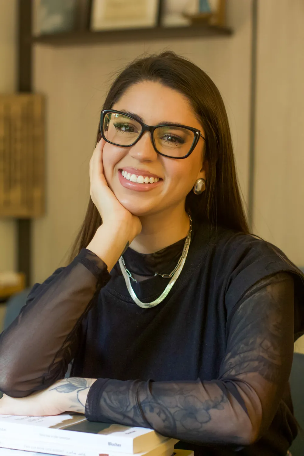 Amanda Anzolim sorrindo em seu consultório, apoiada na mesa ao lado de livros