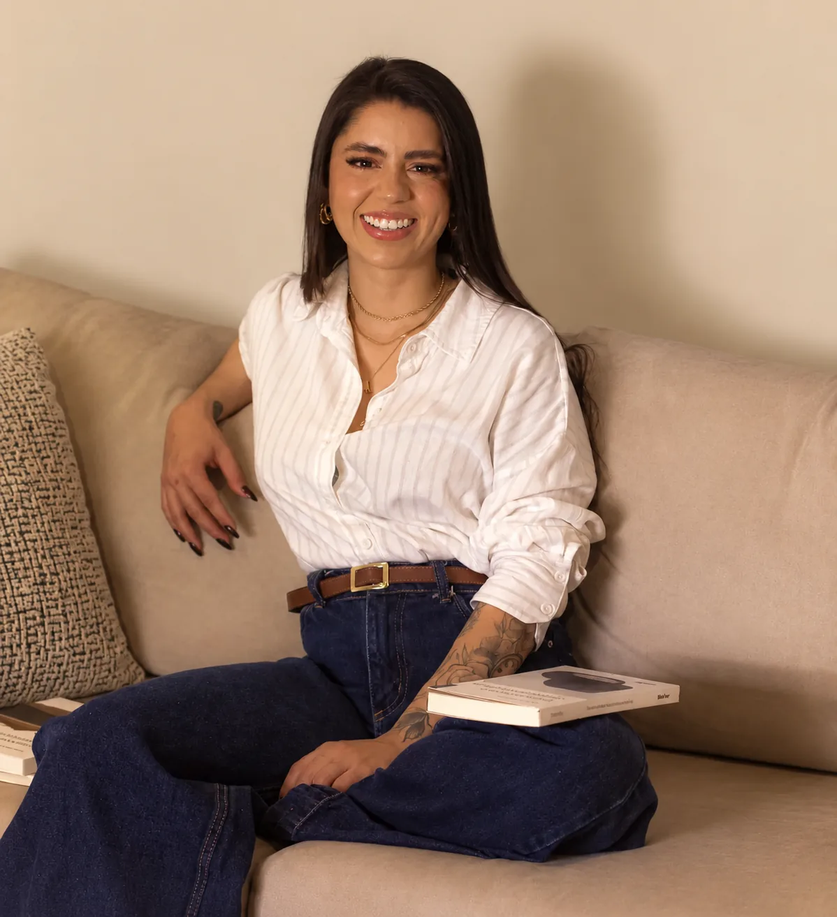 Amanda Anzolim sentada em um sofá bege, sorrindo, com um livro no colo